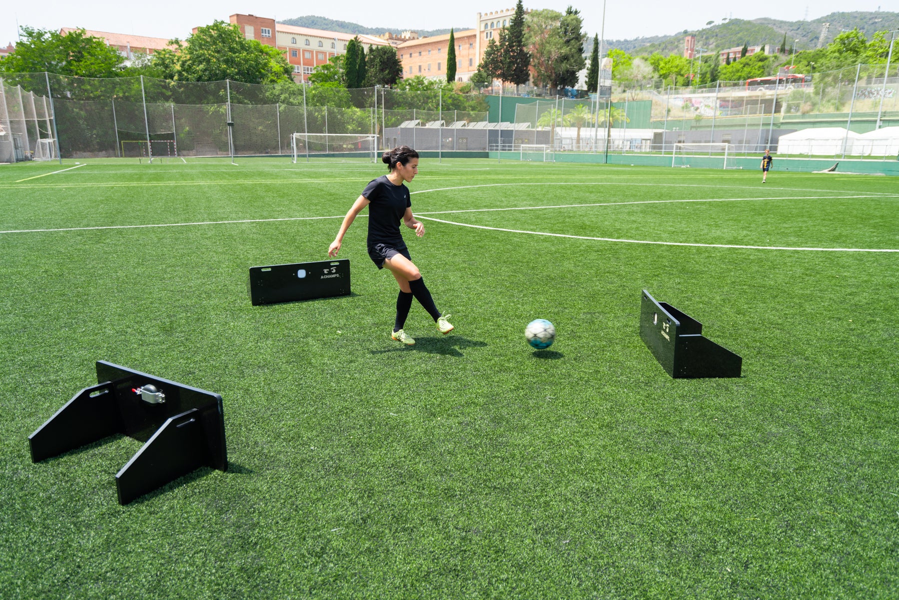 A woman is actively playing soccer on a lush green artificial turf field, dribbling a soccer ball with focus and intent. There are three rebounders on the field with a ROX on each of them, suggesting a training drill setup. The background features a clear sky and a hilly landscape with trees and buildings, indicating the field is located in a scenic, urban area.