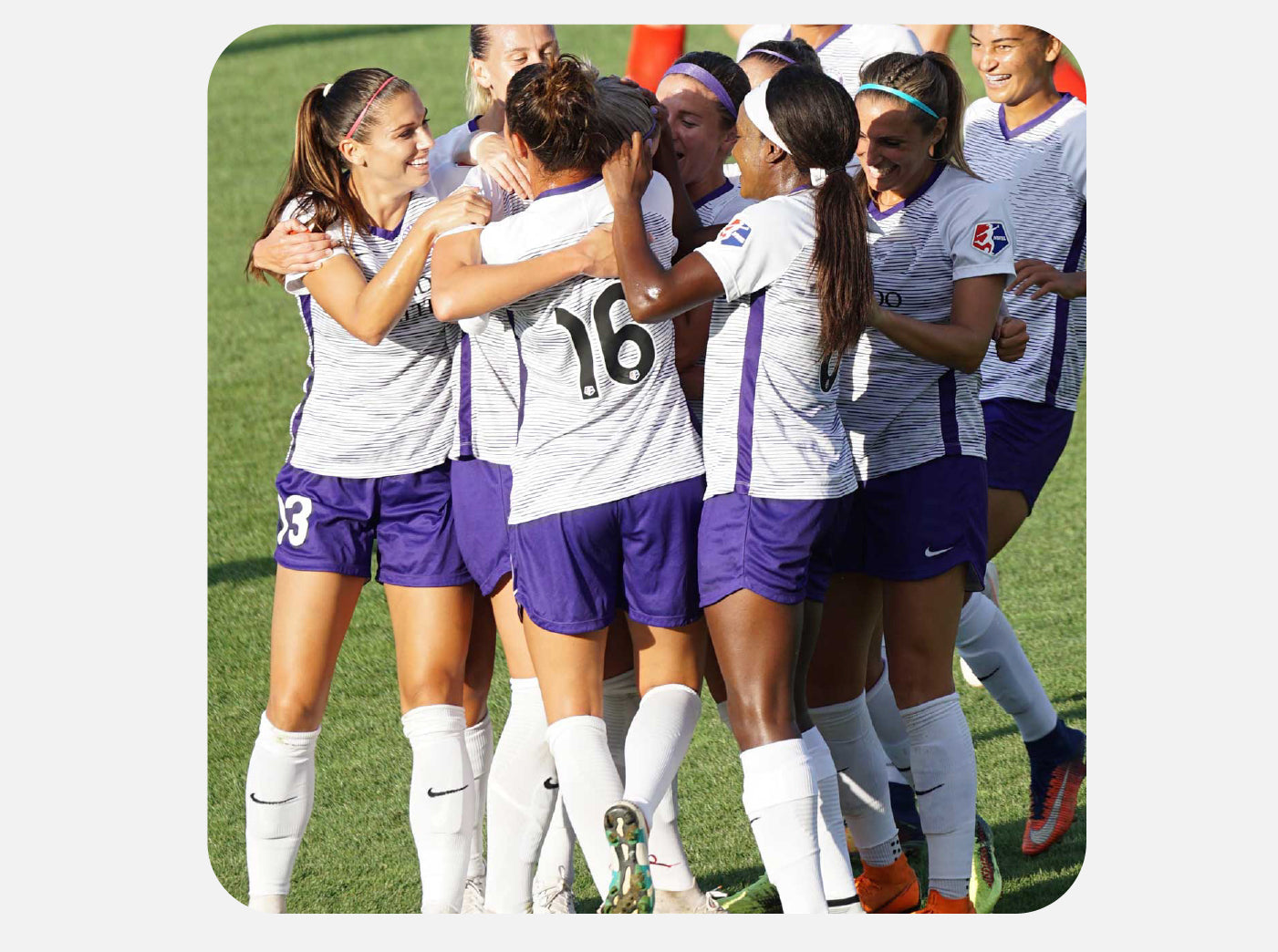 A group of female soccer players in matching white and purple uniforms are joyfully embracing each other on the field in a celebratory huddle. Their expressions are one of happiness and triumph, likely following a successful play or victory in their game. They are wearing white soccer socks and various colored soccer cleats, and the sun is shining down on them, highlighting the camaraderie and athletic spirit of the moment.
