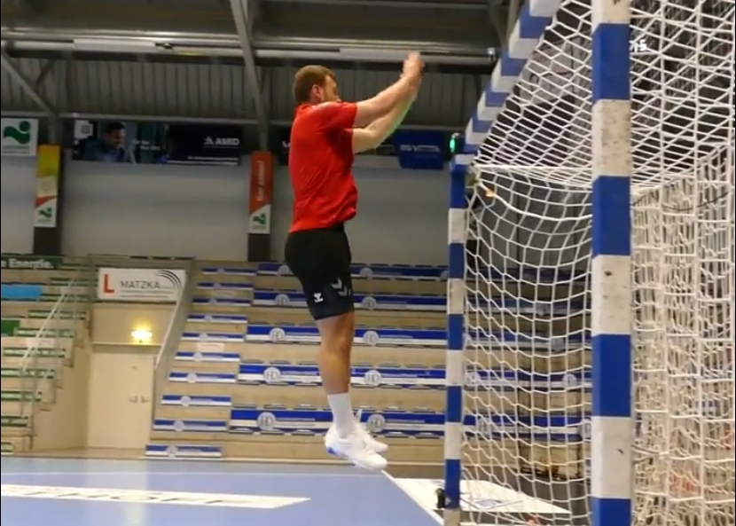 handball player in red shirt and black shorts jumping over a blue and white net in an indoor sports facility.