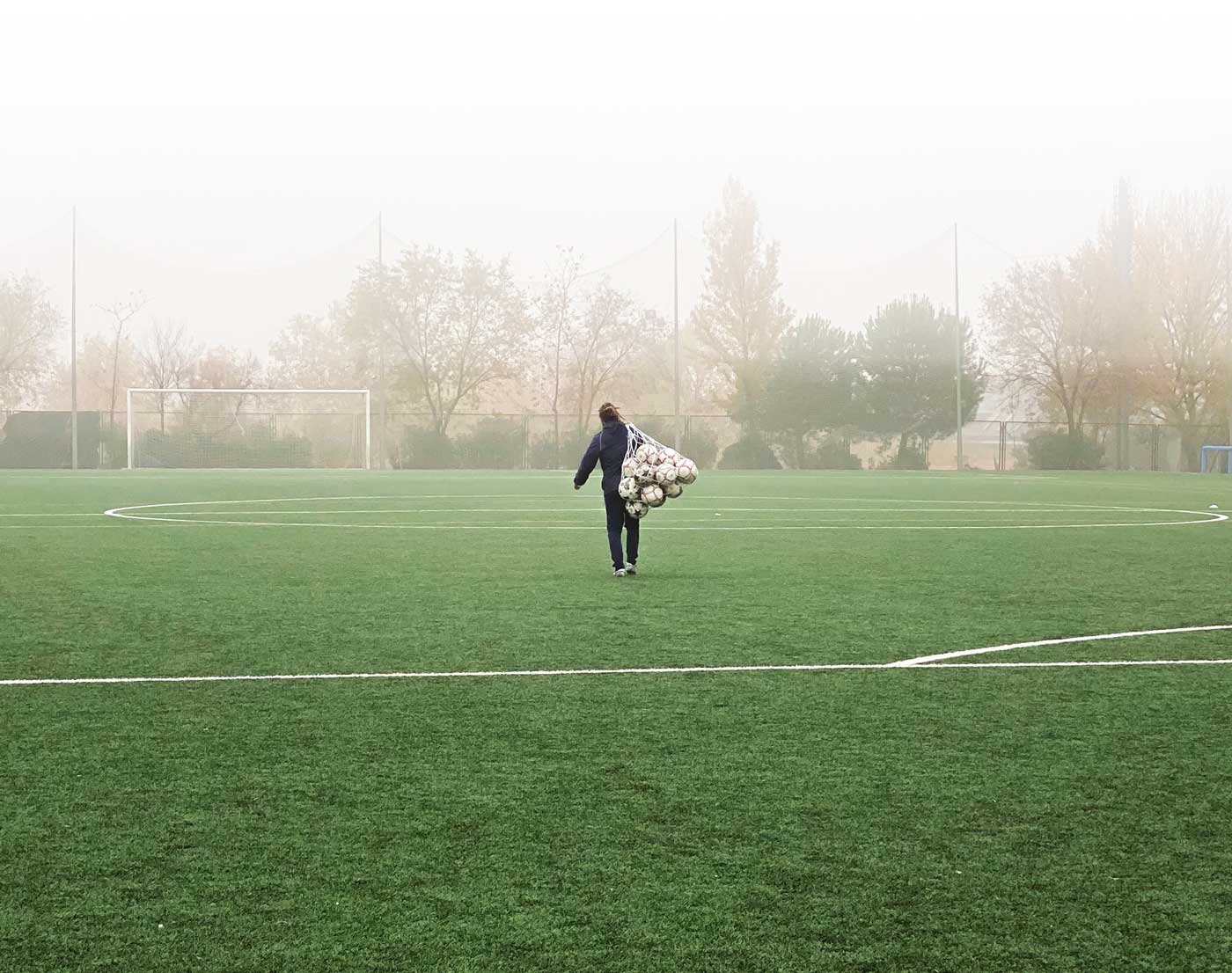 A Soccer trainer is seen walking across a foggy soccer field, carrying a large net bag full of soccer balls over their shoulder. The soccer goal is visible in the background, partially obscured by the mist, and the trees lining the field are faintly visible through the fog. By the leaning of the trainer we can assume He is struggling in carrying that bag of soccer balls all alone.
